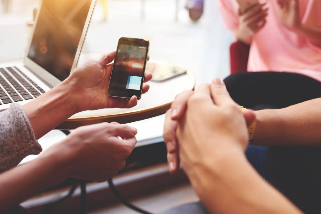 Closely of hand is holding cell phone with copy space screen for your advertising content. Two person are watching video in internet via mobile phone while are sitting near open net-book on the table